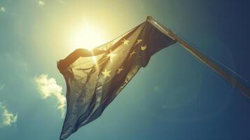 A flag is waving proudly as sunlight pours through its fabric, creating a vibrant display against the backdrop of a mostly clear blue sky with a few scattered clouds. photo