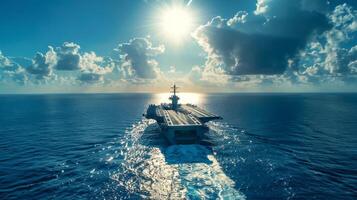 An aircraft carrier moves steadily across the vast blue ocean, illuminated by the midday sun, while fluffy clouds drift in the background, creating a serene maritime view. photo