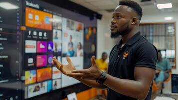 A young man presents confidently to an audience, gesturing towards a large digital screen displaying various colorful applications and content in a contemporary office setting. photo