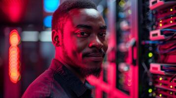 A young man smiles while standing next to high tech server racks illuminated by colorful LED lights, showcasing a modern data center environment during the evening. photo