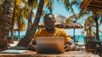 A man sits at a wooden table on a beach, focused on his laptop while wearing sunglasses. Palm trees sway gently in the breeze, creating a tropical atmosphere by the ocean. photo