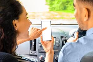 Young mixed race couple having car trip together, using mobile application on smartphone, shot from back seat, closeup. Brunette lady pointing at cellphone with empty screen, mockup photo