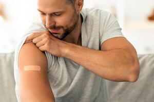 Covid-19 vaccination concept. Young cheerful caucasian man showing shoulder with medical plaster after vaccination, sitting on sofa at home, empty space photo