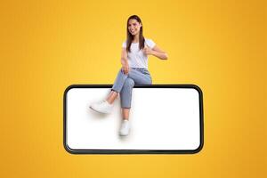 Positive casual millennial lady sitting on big horizontal smartphone with blank screen and showing thumbs up ok gesture, recommending newest mobile application, isolated on orange studio background photo