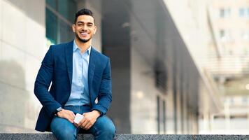 Cheerful middle eastern millennial man in suit IT manager or businessman sitting by modern office building, holding mobile phone, smiling at camera, panorama with copy space. Modern business photo