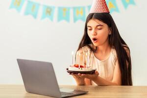 Indian teenage girl in festive hat having online birthday party, blowing candles on cake in front of laptop webcam. Happy adolescent celebrating holiday on internet, chatting with family on web photo
