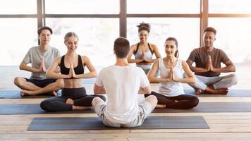 Group of peaceful people practicing yoga with coach in gym studio, making meditation exercises with instructor, panorama photo