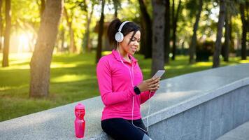 Black girl jogger using sport app for smartphone while resting during training in public park, empty space photo