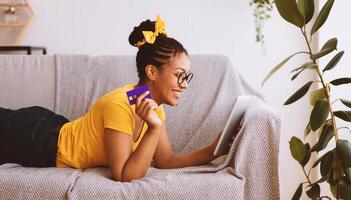 Online Shopping Concept. Happy Black Woman In Glasses Holding And Using Digital Tablet And Credit Card, Buying And Ordering Things In Internet Shop Lying On The Couch. Sale, Retail And Purchase photo