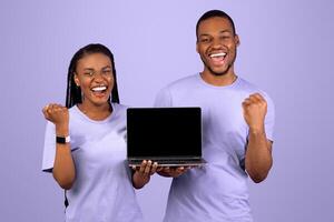 Yes. Portrait of excited happy black couple holding pc laptop with blank screen shaking clenched fists making winner gesture, showing device with empty free space for mock up, violet studio background photo