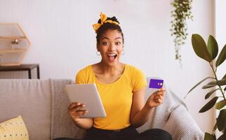 Overjoyed african girl showing credit card, using tablet, sitting on sofa, copy space photo