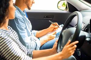 Handsome arab man instructor examinating brunette lady student, taking notes at chart while sitting by smiling young woman driving auto and waiting for test results, side view, copy space photo