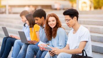 Group Of Students Learning Using Gadgets Sitting On College Steps Outdoor Preparing For End-Of-Semester Exams Together. photo