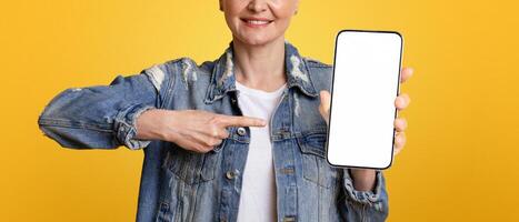 Smartphone Mockup. Smiling Middle Aged Woman Pointing At Black Blank Mobile Screen, Standing Over Yellow Background, Cropped Image, Panorama photo