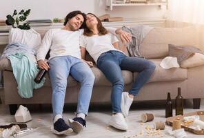 After party. Drunk millennial couple sleeping tegether on couch in messy room with empty beer bottles and box from pizza around photo