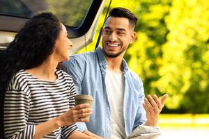 Happy lovers travelling by car together, having coffee break at country side, sitting on open auto trunk, having conversation and drinking tea, looking at each other, cheerfully smiling photo
