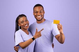 Happy Black Couple Holding And Pointing Finger At Credit Card Recommending Service Smiling To Camera Over Purple Background. Excited Bank Customers Showing Card For Payments Posing In Studio photo