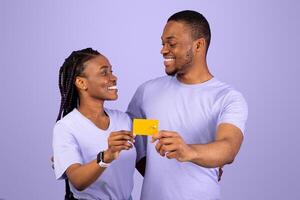 Happy Black Couple Hugging, Holding And Showing Golden Credit Card To Camera, Looking At Each Other, Recommending Banking Service Over Purple Violet Studio Background, Free Copy Space photo