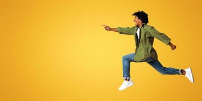 Look There. Overjoyed black guy pointing aside while jumping up in air, cheerful young african american man in motion demonstrating free copy space on beige background, full length shot, panorama photo