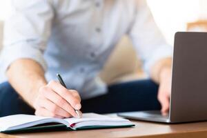 Note Taking. Closeup of unrecognizable male person using laptop, writing in notebook, blurred background photo