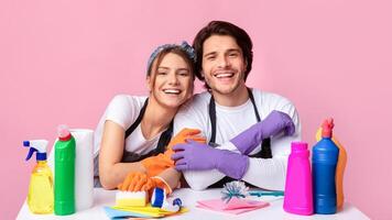 Cheerful Couple Sitting At Table With Lots Of Detergents And Cleaning Supplies, Hugging And Smiling At Camera, Ready To Spring-Clean, Pink Background photo