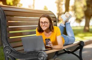 Cheerful Student Girl Using Laptop Computer Browsing Internet Lying On Bench In Park Outside. Online Learning Concept photo