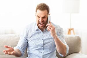 Angry irritated man talking on mobile phone and shouting, sitting on sofa, copyspace photo