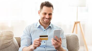 Online Payment. Frontal portrait of man holding debit card and using smartphone for shopping at home, copy space photo