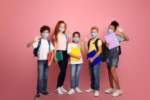 Education during virus pandemic concept. Full length portrait of schoolkids in medical masks waving on pink background photo