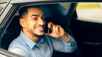 Cheerful middle-eastern young man manager talking to business partner on cellphone while going to office by taxi, sitting at car back seat, closeup portrait, copy space. Businessman lifestyle concept photo