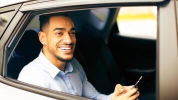 Positive middle-eastern businessman checking emails while going to office by car, sitting on car back seat and using smartphone, closeup photo, shot from outside, side view, copy space photo