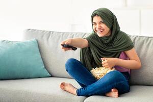 Domestic entertainments. Happy Muslim teenage girl in hijab with TV controller enjoying movie and eating popcorn indoors. Happy adolescent in traditional headscarf watching television on couch at home photo