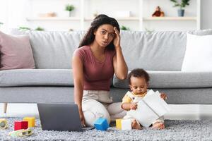 Working At Home Problems. Stressed Mother Trying To Work On Laptop While Baby Distracting Her, Worried African American Woman Having Troubles With Remote Work While Taking Care About Child photo