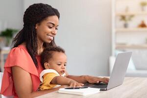 Portrait Of Young Woman With Baby On Hands Using Laptop At Home, Happy Smiling African American Lady Study Or Working Remotely On Computer While Looking After Her Child, Side View, Free Space photo