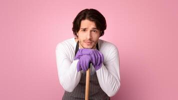 Tired Man Leaning At Mop Handle, Resting Head On Hands Exhausted After Spring Cleaning, Standing Over Pink Background With Free Space photo