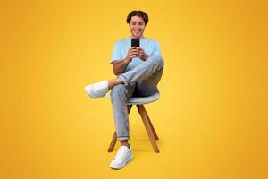 Great App. Contented Casual Man Using Smartphone Checking New Mobile Application, Chatting With Friends Texting Online Sitting In Chair, Orange Wall. Modern Mobile Communication, People And Gadgets photo
