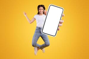 Overjoyed young casual lady showing empty smart phone screen while jumping up over yellow studio background, collage, full size photo. Happy woman recommending new mobile app, free copy space, macro photo