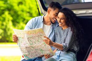 Happy young lovers cuddling while sitting on auto trunk and watching road map, copy space. Handsome arab guy bonding with his beautiful girlfriend, having break while car trip together photo