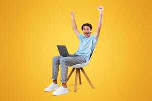 Win. Vertical portrait of excited guy celebrating success shaking clenched fists and raising arms up sitting on chair using laptop computer, reading great news, screaming yes, yellow studio wall photo