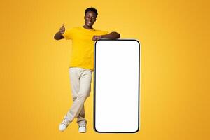 Cheerful African American Guy Leaning At Big Smartphone With White Blank Screen And Showing Thumb Up, Smiling Black Man Recommending New Mobile App While Standing Over Beige Background, Mockup Image photo