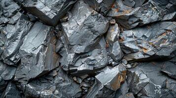 Jagged dark gray rocks are stacked closely together, revealing intricate textures and patterns, with occasional orange streaks adding contrast, illuminated by natural light. photo