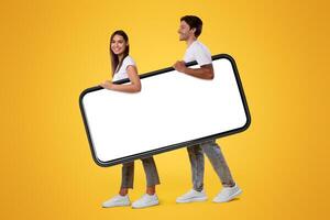 Millennial couple holding big huge smartphone with empty screen on orange studio background. Mockup for advertisement, mobile app or new website promo. Cell phone display template, free copy space photo