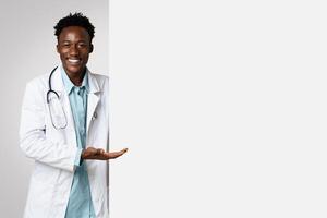 Smiling black male doctor pointing at big blank advertisement board with open palm, professional therapist demonstrating empty copy space for text or vaccination campaign design, panorama photo