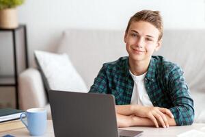 Smiling teen guy studying at home, using laptop, making homework photo