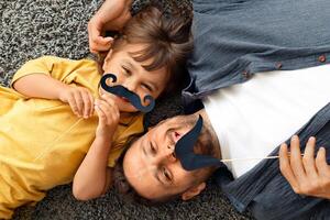 Family fun. Top view portrait of positive father and little boy smiling to camera with moustache props, lying on grey carpet at home photo