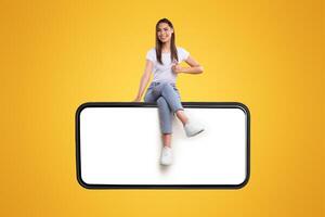 Cheerful young woman sitting on big huge horizontal new cellphone with white empty screen and showing thumb up gesture, mockup, casual lady enjoying newest entertaining mobile app, orange wall photo