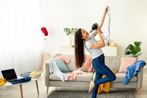 Indian teenage girl dancing and singing in messy room, using blowdryer as microphone, giving domestic concert. Cute adolescent lady having fun, entertaining herself during covid lockdown photo
