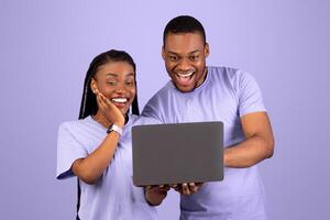 Portrait of happy amazed African American couple holding and using laptop, emotional black guy showing lady something exciting content, pointing at notebook screen or typing on keyboard, copy space photo