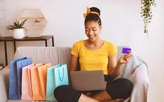 Online Payment Concept. Smiling african american lady holding credit card and using laptop, ordering goods and purchases via internet, sitting on the sofa with colorful shopping bags in living room photo