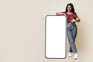 Stylish millennial black woman pointing at big mobile phone with white blank display, mockup, happy african american lady in casual recommending newest mobile application, beige studio background photo